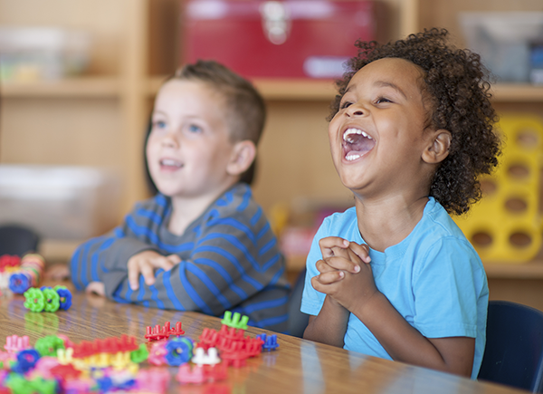 Preschool children are playing with toy blocks and laughing together at a classroom table.